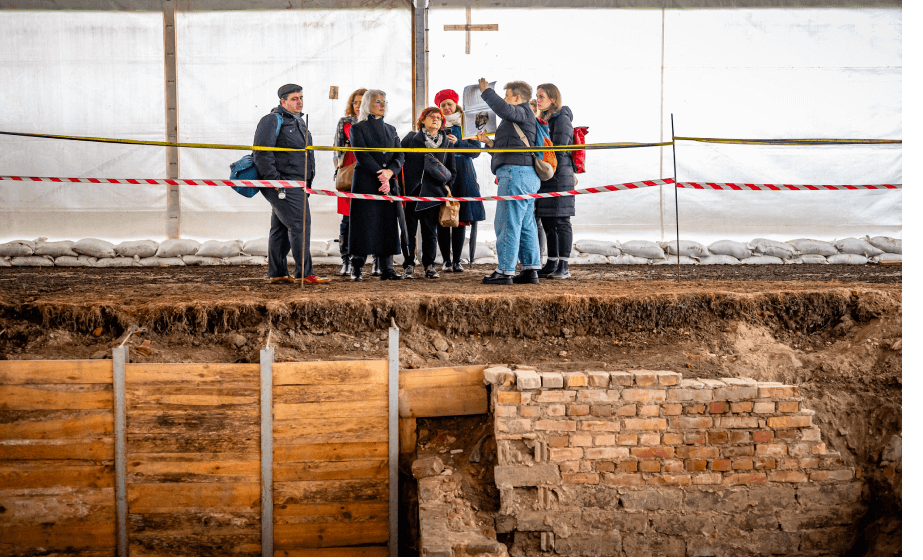 A group of people stand at the edge of an excavation site. Behind them is plastic tarpaulin and in front of them is red and white tape marking off the excavation site. A member of the group holds up a book while the others looks on. Brickwork and wooden planks are visible in the excavation site.