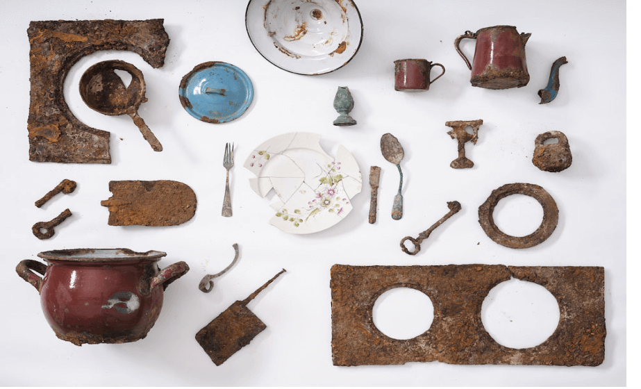 A selection of objects found in the excavation site of the Warsaw ghetto. The objects are laid out on a white background and is taken from above. In the centre of the image is a white plate with a flowery pattern that has been pieced back together and is missing some parts. Surrounding the plate are pots, bowls, and a teapot among other things.