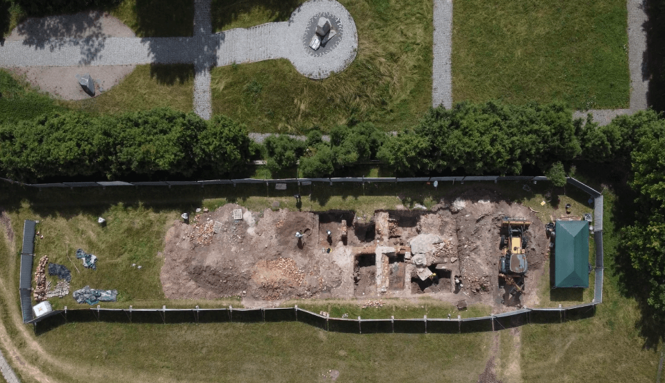 An aerial photo of the Warsaw Ghetto site shows an excavation site composed of a number of rectangular holes surrounded by an iron fence. There is a small green shed and a yellow digger within the fenced area. Above the excavation site there is a grassy mound with steps leading up to a large stone monument.